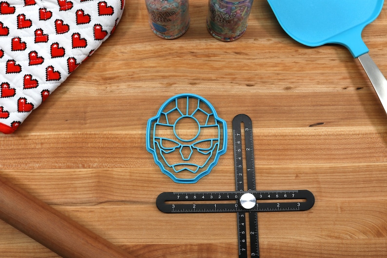 May include: A blue plastic cookie cutter shaped like a cartoon face with a large circle in the center of the face. The cookie cutter is on a wooden surface with a black ruler and a rolling pin.