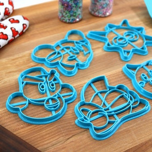 May include: A set of six blue plastic cookie cutters shaped like cartoon characters. The cutters are on a wooden surface.