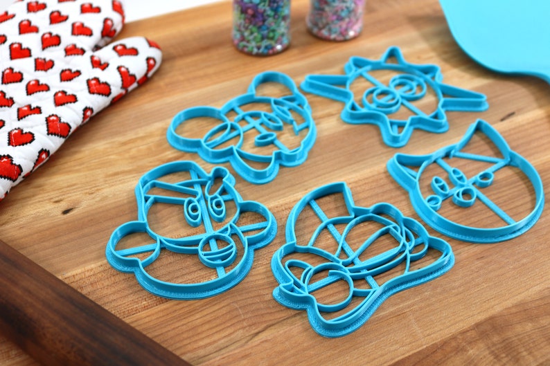 May include: A set of six blue plastic cookie cutters shaped like cartoon characters. The cutters are on a wooden surface.