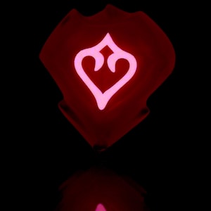 May include: A red, heart-shaped object with a pink, stylized heart design inside. The object is glowing and reflecting on a black surface.