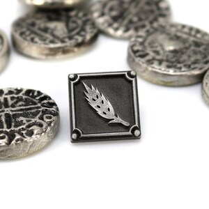May include: A silver metal pin with a black square background and a silver wheat stalk design.