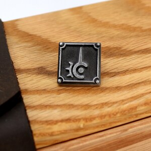 May include: A silver metal pin with a black background and a stylized sword and circle design. The pin is on a wooden surface.
