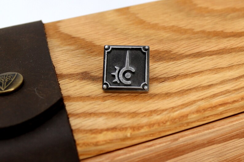 May include: A silver metal pin with a black background and a stylized sword and circle design. The pin is on a wooden surface.