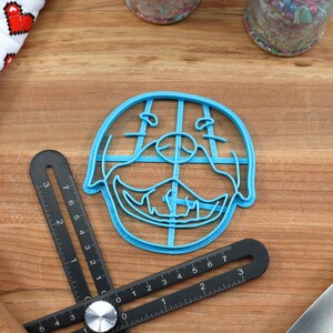 May include: A blue plastic cookie cutter shaped like a dog's face. The cutter has a grid pattern inside the outline of the dog's face.
