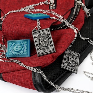 Puede incluir: Tres cadenas plateadas con colgantes que cuelgan de una bolsa roja. Los colgantes son rectangulares y presentan un dise&ntilde;o de cara en blanco y negro. Un colgante es azul.