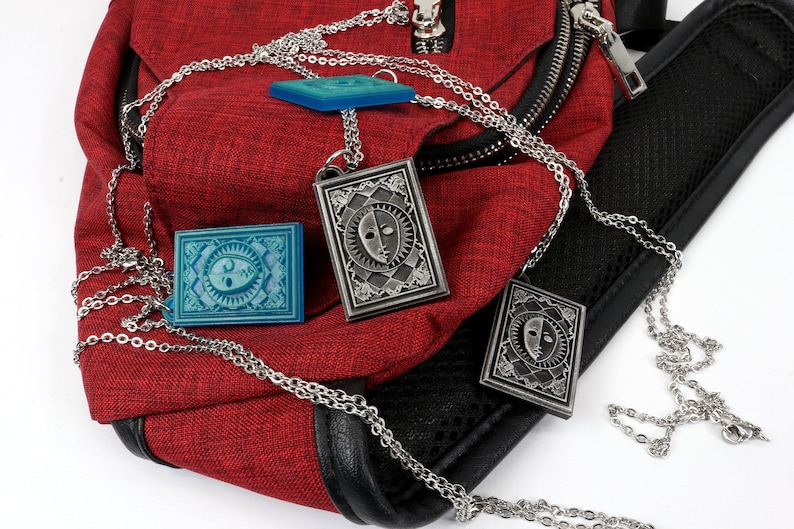 Puede incluir: Tres cadenas plateadas con colgantes que cuelgan de una bolsa roja. Los colgantes son rectangulares y presentan un dise&ntilde;o de cara en blanco y negro. Un colgante es azul.