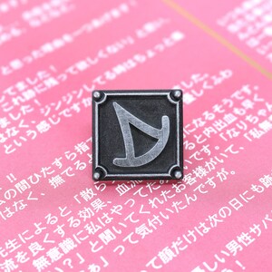 May include: A silver metal pin with a black background and a stylized silver letter D in the center. The pin is on a pink surface with Japanese text.