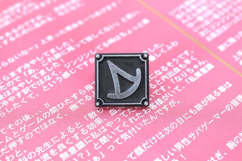 May include: A silver metal pin with a black background and a stylized silver letter D in the center. The pin is on a pink surface with Japanese text.