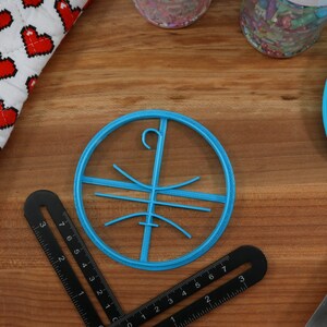 Op de afbeelding: Een ronde koekjesvormer van blauw plastic met een haak aan de bovenkant en een ontwerp van lijnen erin. De vormer ligt op een houten oppervlak.