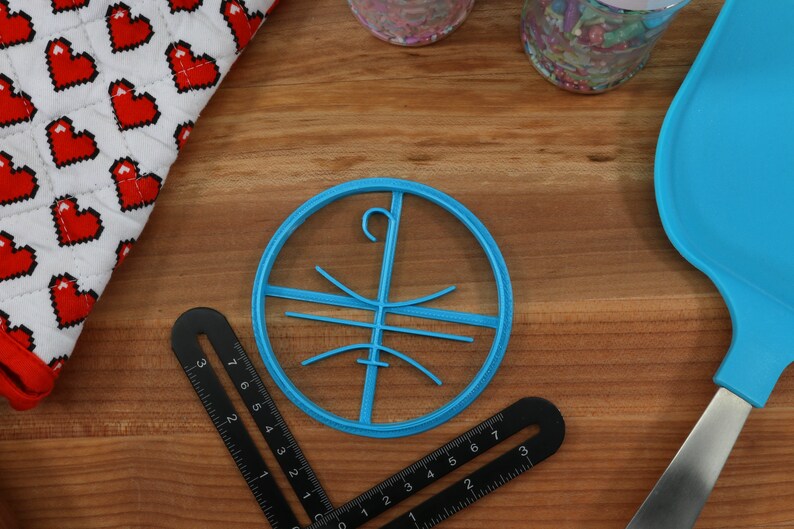 Op de afbeelding: Een ronde koekjesvormer van blauw plastic met een haak aan de bovenkant en een ontwerp van lijnen erin. De vormer ligt op een houten oppervlak.