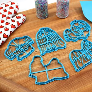May include: A set of five blue plastic cookie cutters shaped like Shih Tzu dogs. The cutters are on a wooden surface with a rolling pin, a red oven mitt, and two jars of sprinkles.