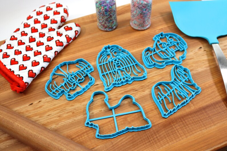 May include: A set of five blue plastic cookie cutters shaped like Shih Tzu dogs. The cutters are on a wooden surface with a rolling pin, a red oven mitt, and two jars of sprinkles.
