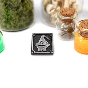 May include: A silver metal pin with a black background featuring a stylized image of a potion bottle with a cork stopper.