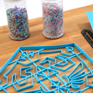 May include: A blue plastic cookie cutter shaped like a shield with a design inside. Two glass jars filled with colorful sprinkles are in the background.