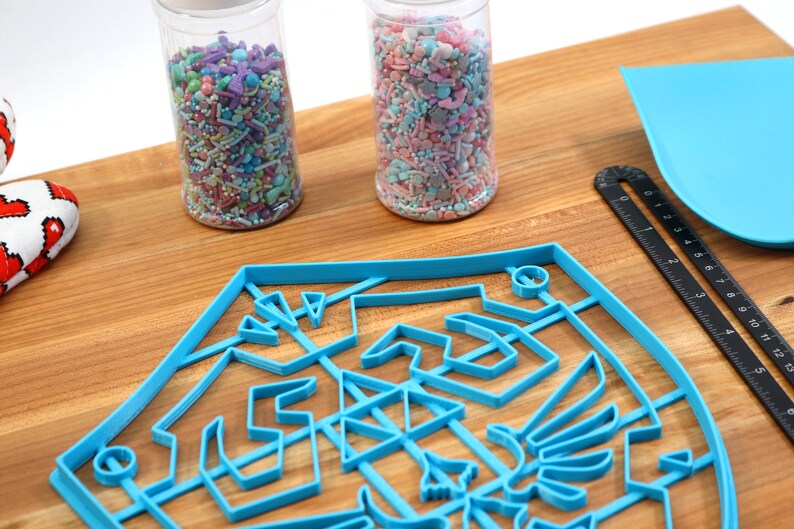 May include: A blue plastic cookie cutter shaped like a shield with a design inside. Two glass jars filled with colorful sprinkles are in the background.