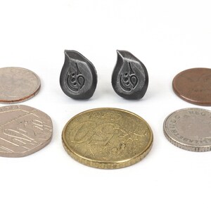Puede incluir: Dos peque&ntilde;os pendientes de color plateado en forma de l&aacute;grima con un dise&ntilde;o estilizado. Los pendientes se muestran junto a varias monedas para comparar el tama&ntilde;o.