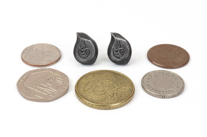 Puede incluir: Dos peque&ntilde;os pendientes de color plateado en forma de l&aacute;grima con un dise&ntilde;o estilizado. Los pendientes se muestran junto a varias monedas para comparar el tama&ntilde;o.