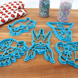 May include: A collection of blue plastic cookie cutters in various cartoon character shapes, including a crown and a rabbit. A red and white heart-patterned oven mitt and sprinkles are also visible.