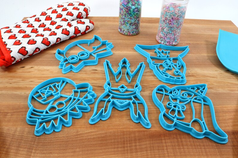 May include: A collection of blue plastic cookie cutters in various cartoon character shapes, including a crown and a rabbit. A red and white heart-patterned oven mitt and sprinkles are also visible.