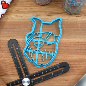 May include: Blue plastic cookie cutter shaped like a dog wearing sunglasses. The cookie cutter is on a wooden surface.