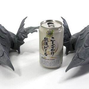 Op de afbeelding: Twee grijze 3D-geprinte drakenkoppen met vleugels die elkaar aankijken, met een blikje Japans bier in het midden. Het blikje heeft Japanse tekst en de woorden "Koshihikari Rice" en "Echigo" erop.