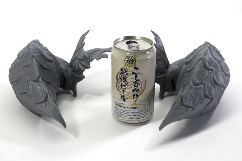 Op de afbeelding: Twee grijze 3D-geprinte drakenkoppen met vleugels die elkaar aankijken, met een blikje Japans bier in het midden. Het blikje heeft Japanse tekst en de woorden "Koshihikari Rice" en "Echigo" erop.