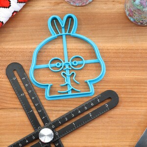 May include: Blue plastic cookie cutter shaped like a cartoon bunny with glasses. The bunny has a wide smile and its ears are sticking up.