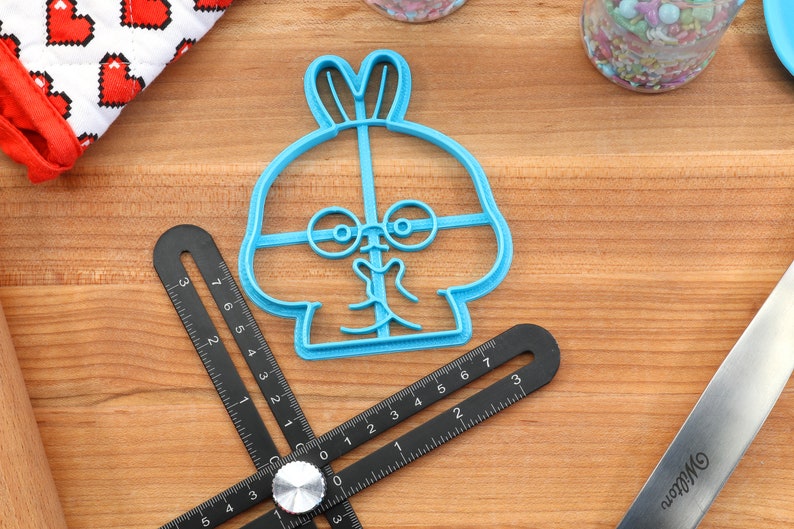 May include: Blue plastic cookie cutter shaped like a cartoon bunny with glasses. The bunny has a wide smile and its ears are sticking up.