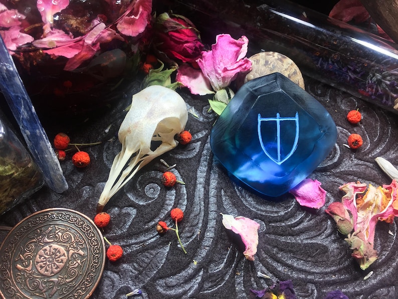 May include: A blue glass shield with a white symbol in the center, surrounded by dried flowers and other natural elements.