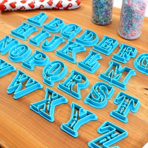 May include: A set of 26 blue plastic alphabet cookie cutters on a wooden surface. The cutters are in a serif font style.