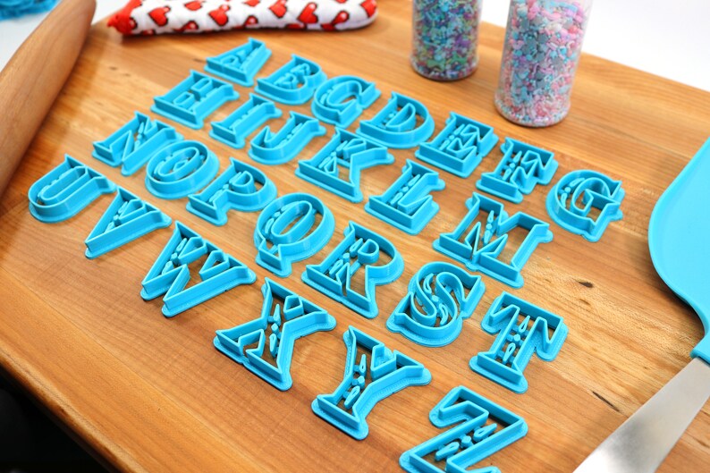 May include: A set of 26 blue plastic alphabet cookie cutters on a wooden surface. The cutters are in a serif font style.