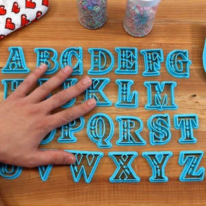 May include: A set of 26 blue plastic alphabet cookie cutters on a wooden surface. The cutters are arranged in alphabetical order and have a decorative design.