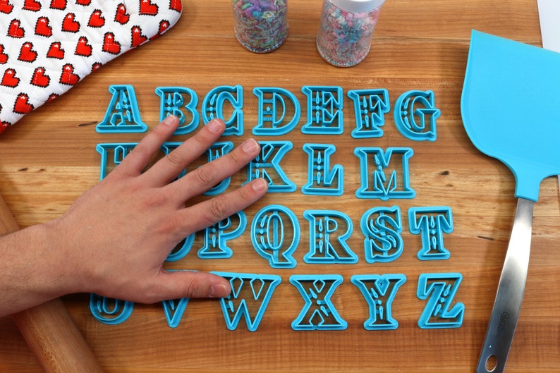 May include: A set of 26 blue plastic alphabet cookie cutters on a wooden surface. The cutters are arranged in alphabetical order and have a decorative design.