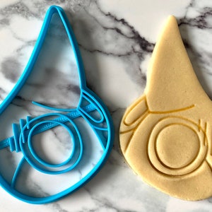 Pode incluir: Um cortador de biscoitos de pl&aacute;stico azul em forma de m&aacute;scara facial, ao lado de um biscoito que foi cortado usando o cortador.