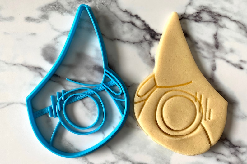 Pode incluir: Um cortador de biscoitos de pl&aacute;stico azul em forma de m&aacute;scara facial, ao lado de um biscoito que foi cortado usando o cortador.