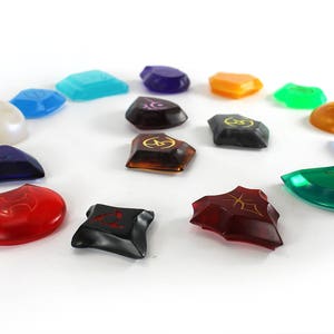May include: A set of 14 colorful, translucent, and opaque gemstone-shaped game pieces with various symbols and designs. The pieces are arranged in a circle on a white background.