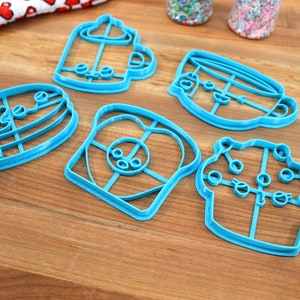 May include: Five blue plastic cookie cutters shaped like a coffee cup, a cupcake, a slice of bread, a pancake, and a mug. The cutters are on a wooden surface.