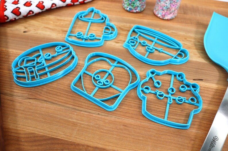 May include: Five blue plastic cookie cutters shaped like a coffee cup, a cupcake, a slice of bread, a pancake, and a mug. The cutters are on a wooden surface.