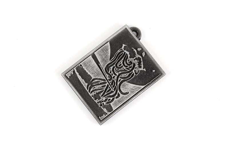 May include: A silver-toned metal pendant with a black and white image of a creature with tentacles. The pendant has a loop at the top for attaching a chain.