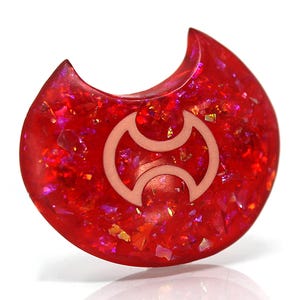 May include: A red and pink resin guitar pick with a white symbol in the center. The pick is shaped like a crescent moon.
