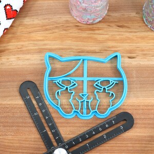 May include: Blue plastic cookie cutter shaped like a cat with its middle fingers raised. The cat has a white outline and a black nose.