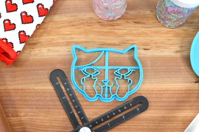 May include: Blue plastic cookie cutter shaped like a cat with its middle fingers raised. The cat has a white outline and a black nose.