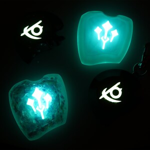 May include: Four glow-in-the-dark resin charms with different designs. Two charms feature a stylized eye symbol, and two charms feature a stylized flower symbol. The charms are a light blue-green color.