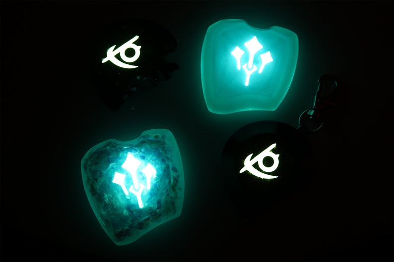 May include: Four glow-in-the-dark resin charms with different designs. Two charms feature a stylized eye symbol, and two charms feature a stylized flower symbol. The charms are a light blue-green color.