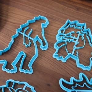 May include: Four blue plastic dinosaur cookie cutters on a wooden surface. The cutters are in the shapes of a Tyrannosaurus Rex, a Triceratops, a Stegosaurus, and a Brachiosaurus.
