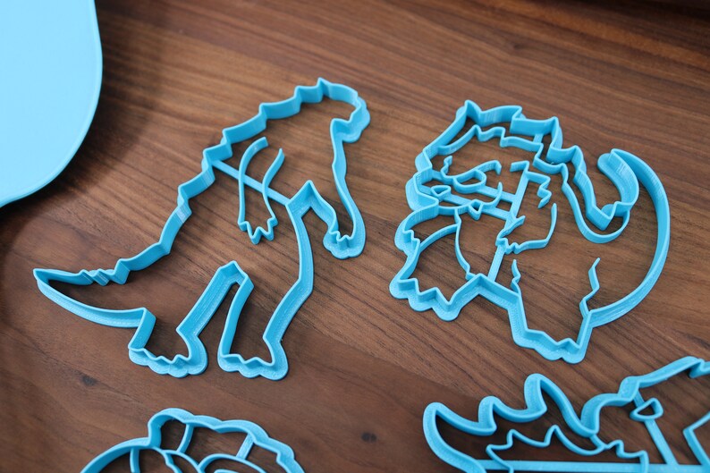 May include: Four blue plastic dinosaur cookie cutters on a wooden surface. The cutters are in the shapes of a Tyrannosaurus Rex, a Triceratops, a Stegosaurus, and a Brachiosaurus.