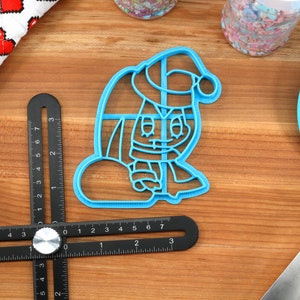 May include: Blue plastic cookie cutter shaped like a cartoon character wearing a Santa hat and holding a bag. The character has a red nose and a white beard.