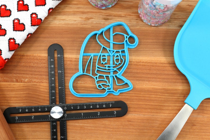 May include: Blue plastic cookie cutter shaped like a cartoon character wearing a Santa hat and holding a bag. The character has a red nose and a white beard.