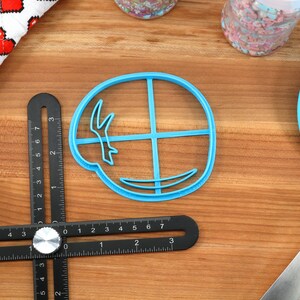 May include: A blue plastic cookie cutter in the shape of a circle with a design inside. A black multi-angle measuring ruler, a blue spatula, and a red and white heart-patterned cloth are also visible on a wooden surface.