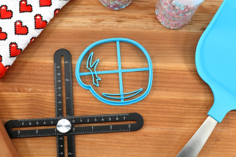 May include: A blue plastic cookie cutter in the shape of a circle with a design inside. A black multi-angle measuring ruler, a blue spatula, and a red and white heart-patterned cloth are also visible on a wooden surface.